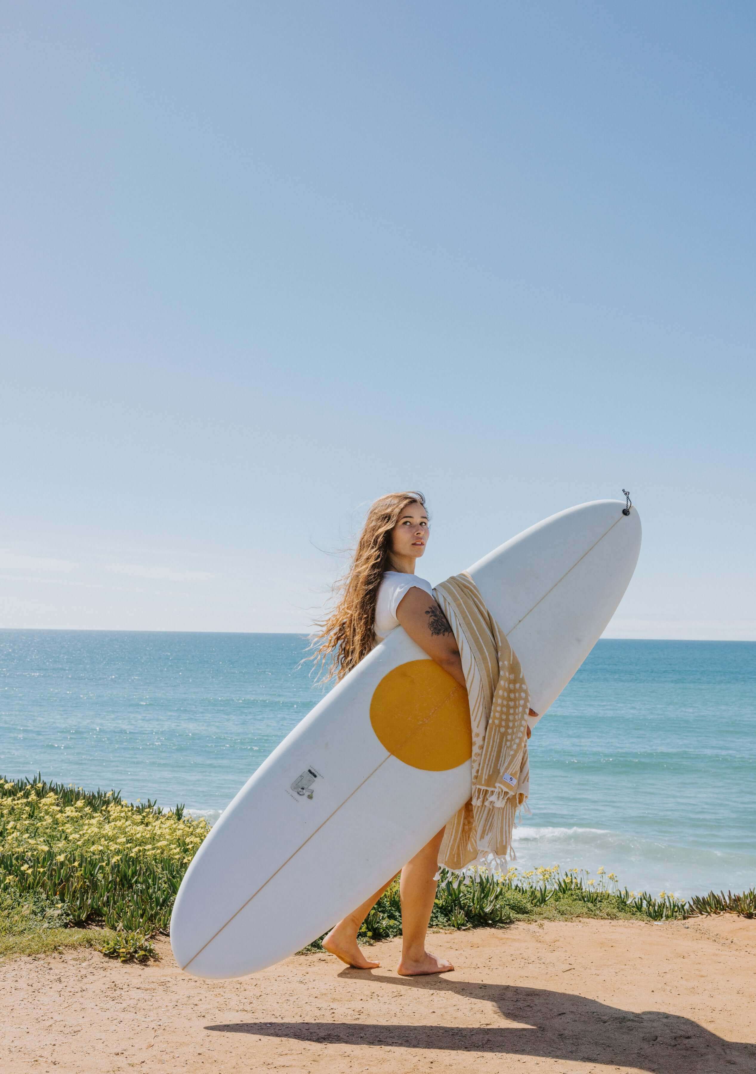 Model above the beach with flowers carrying surfboard and Turkish Honey Beach Towel